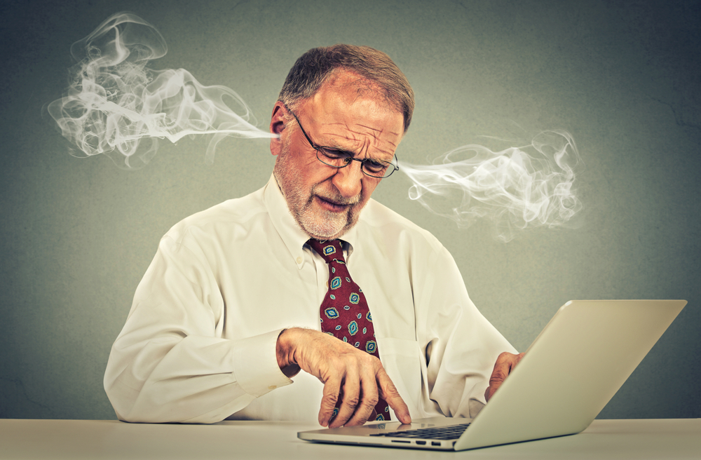 Confused looking elderly mature man with glasses sitting at table working typing on laptop computer